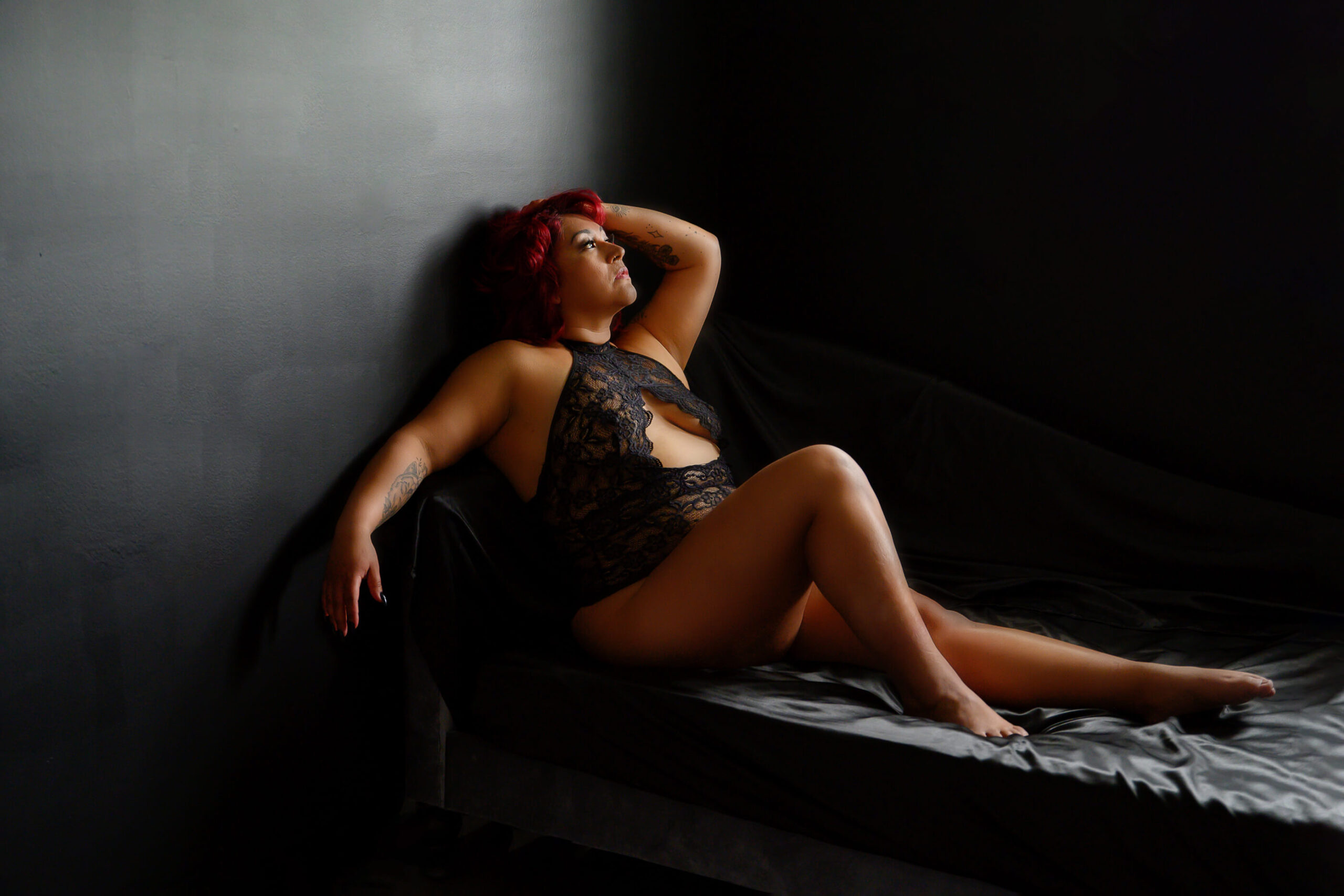 Woman with red hair sitting on a bed with black silk sheets and black wall for her luxury boudoir photography session.