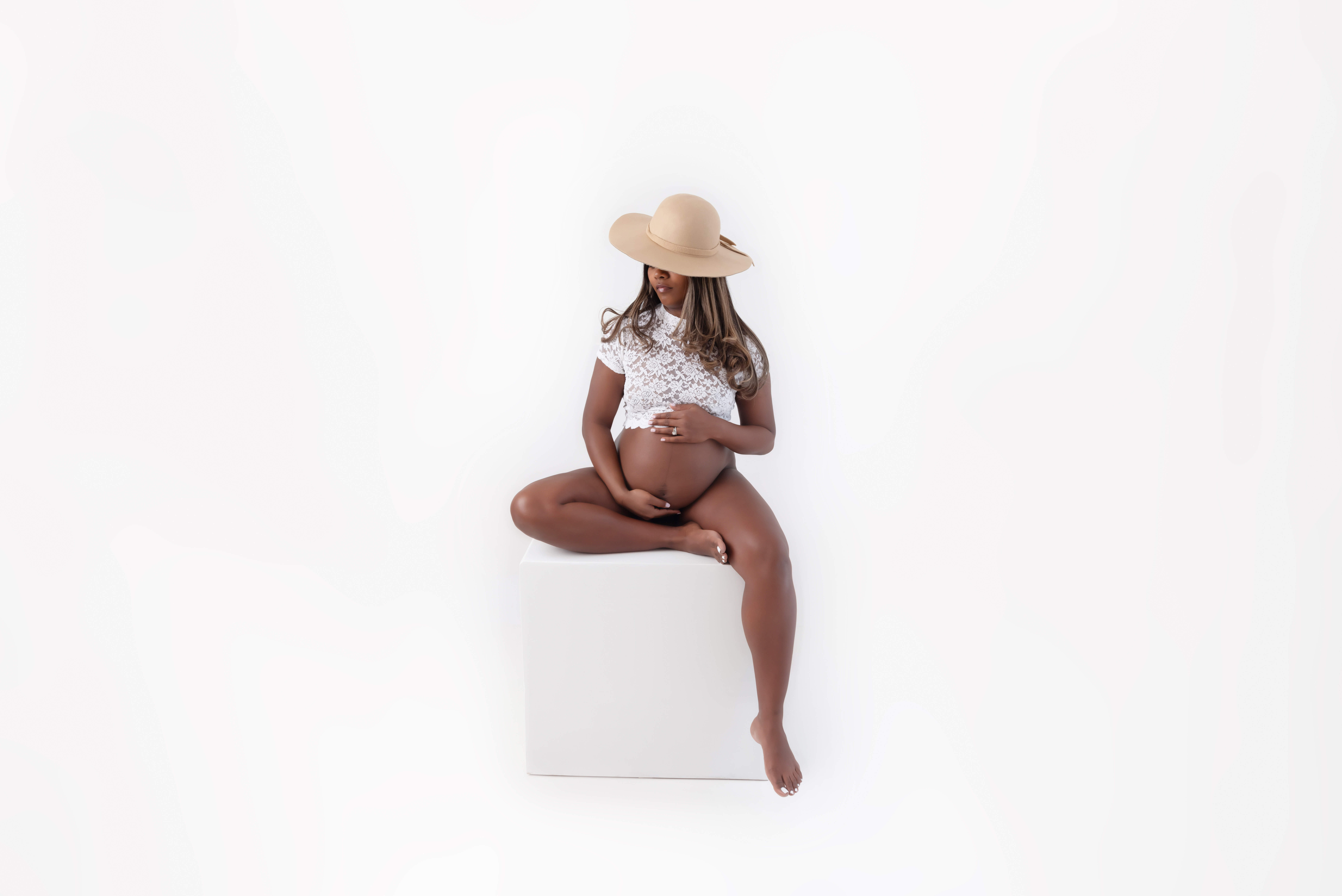 African American pregnant woman wearing a white lace top and beige hat sitting on a white box, for her Maternity Photography Session Toronto Ontario.