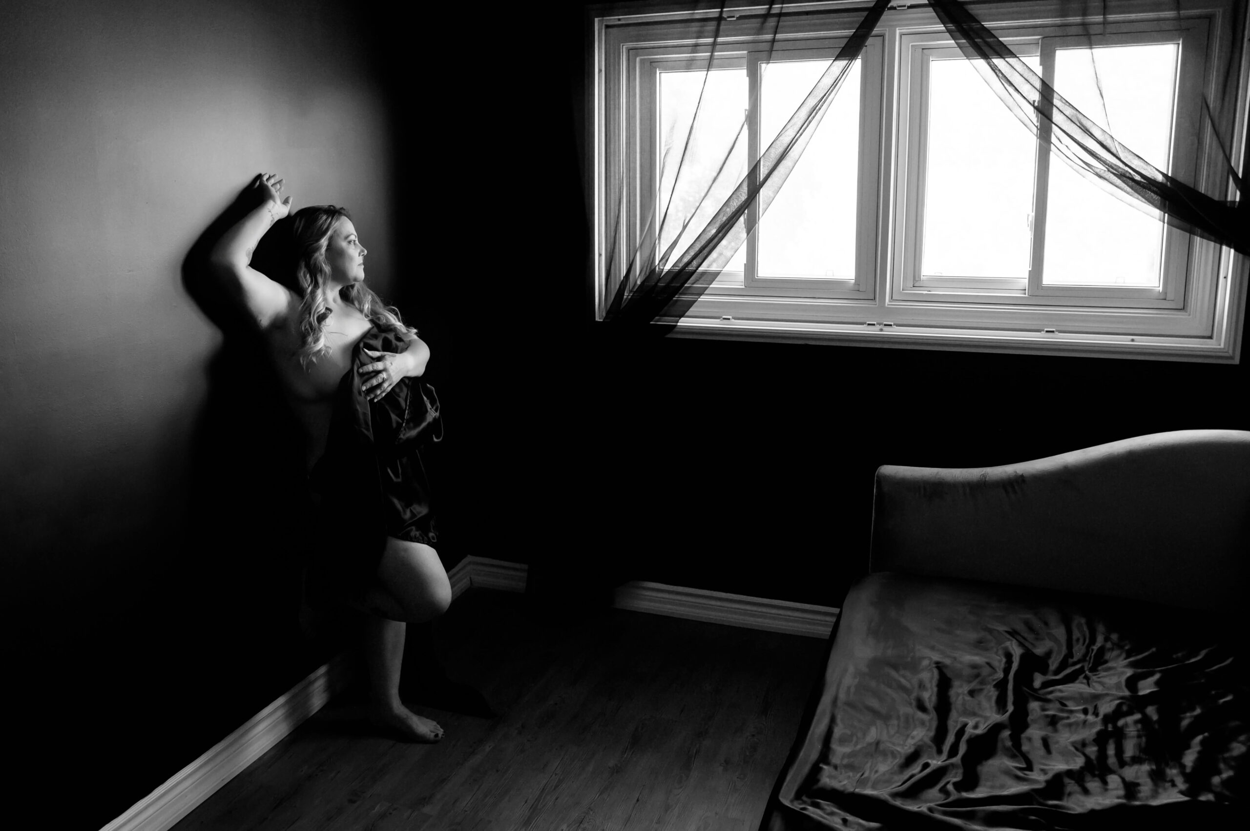 Woman standing against a wall with her arm up looking out the window holding a black silk sheet around her for her luxury boudoir photography session.